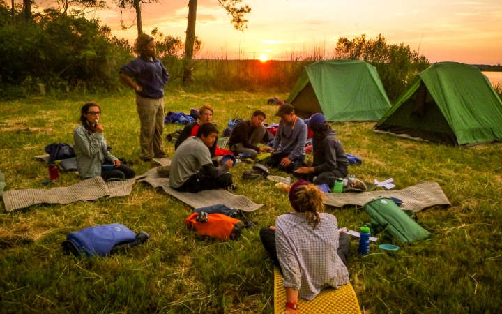 A group of young people rest on pads in the grass near their tents. The sun is setting behind a body of water in the background. 